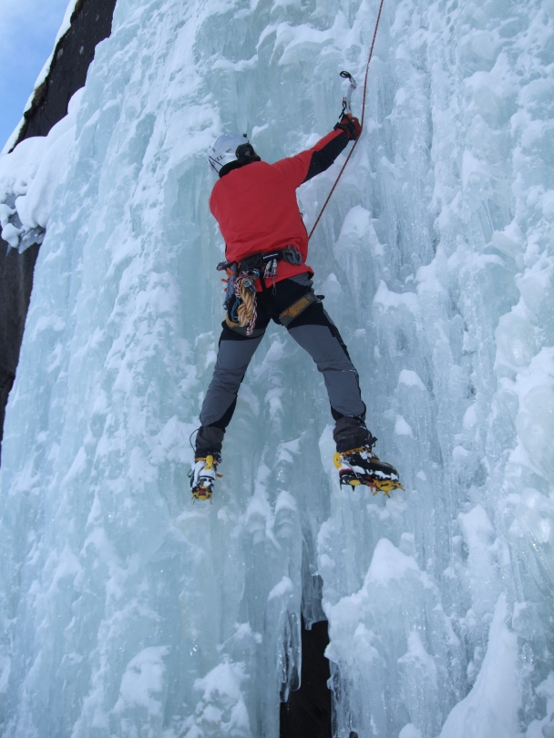 A picture of Jesse ice climbing in Norway. He is using axes and crampons to climb a frozen waterfall. It looks very cold, he is wearing a big red jacket.