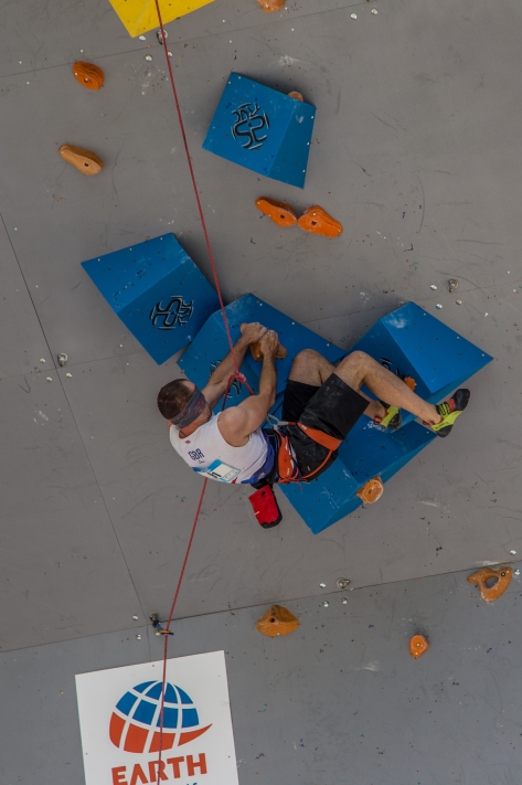 A picture of Jesse climbing through a very steep section on an artificial climbing wall outside. He is climbing over large blue volumes with small orange holds.
