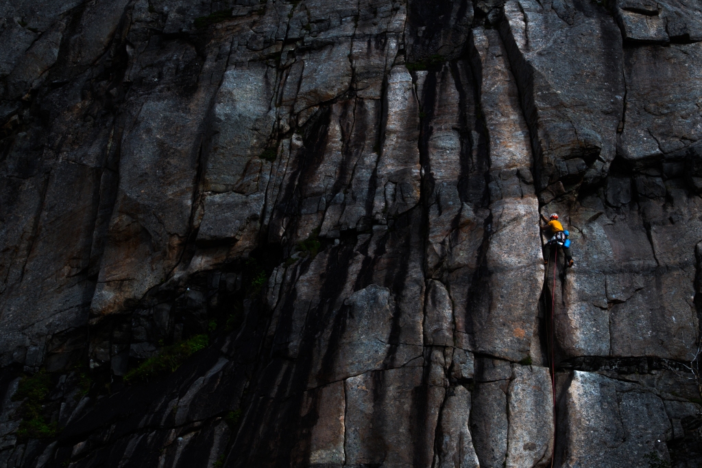 A photo of Jesse climbing Gandalf in Lofoten. The Rock is very dark and he stands out in his orange t-shirt.