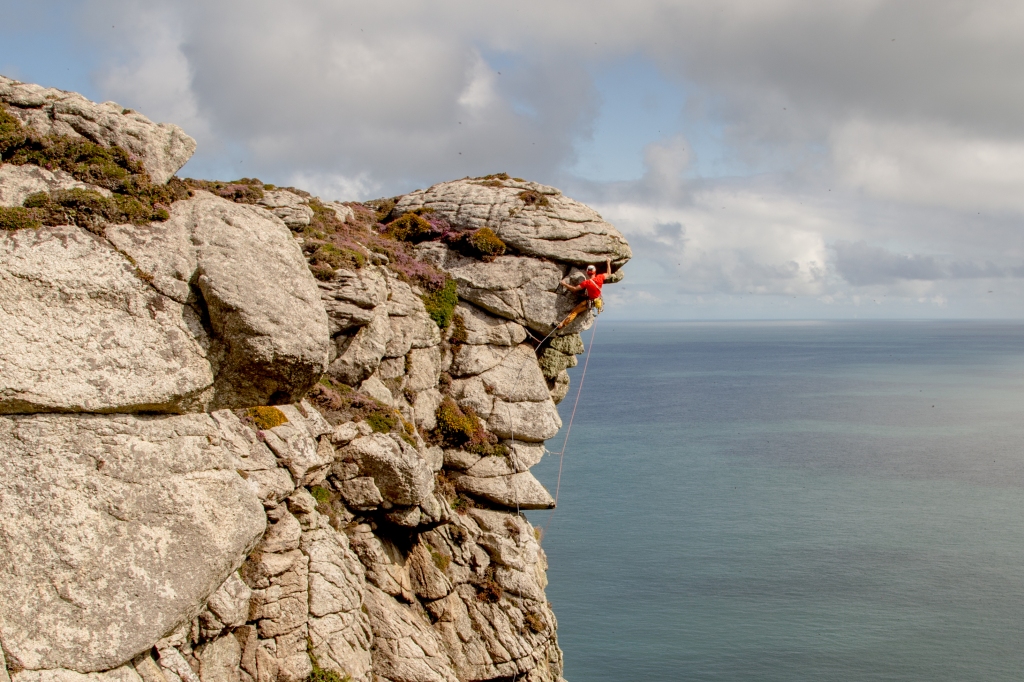 A photo of Jesse nearing the top of a steep overhang that looks out to sea. He is wearing a red T-shirt and is throwing some funky shapes, with the ropes draped below him.