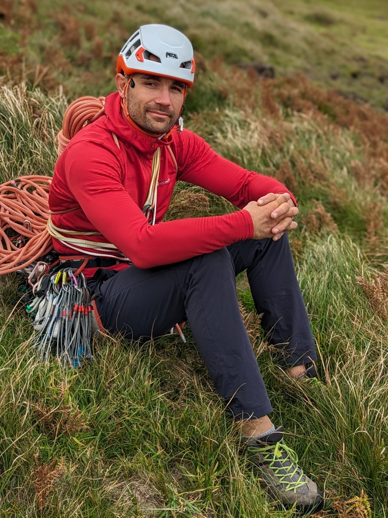 A photo of Jesse pulling a sly smile towards the camera, unaware he is being photographed. He has a coiled rope on his back and is all racked up ready to climb.