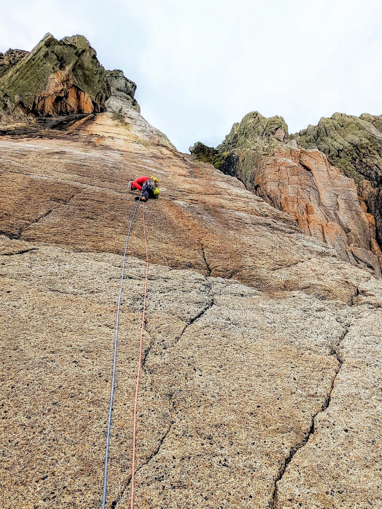 A photo taken from the bottom of Devil's slide, looking up at a huge sweeping slab above, shaped like a slide in a playground but on a giant scale. Jesse is in shot, leading the first pitch. He is trying to fiddle some gear into a horizontal break. 