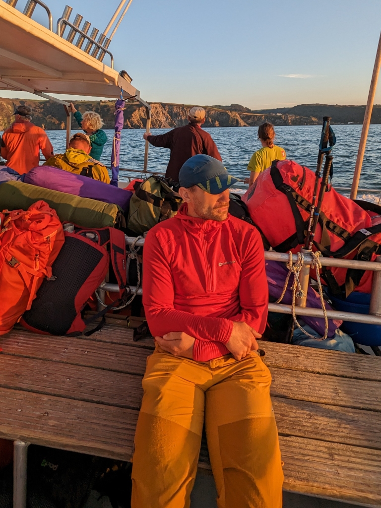 A photo of Jesse sat on a wooden bench on the dive boat. The sun is setting and the photo has an orange glow. 5 people are in the background looking out towards the north coast of Devon, which has very steep cliffs.